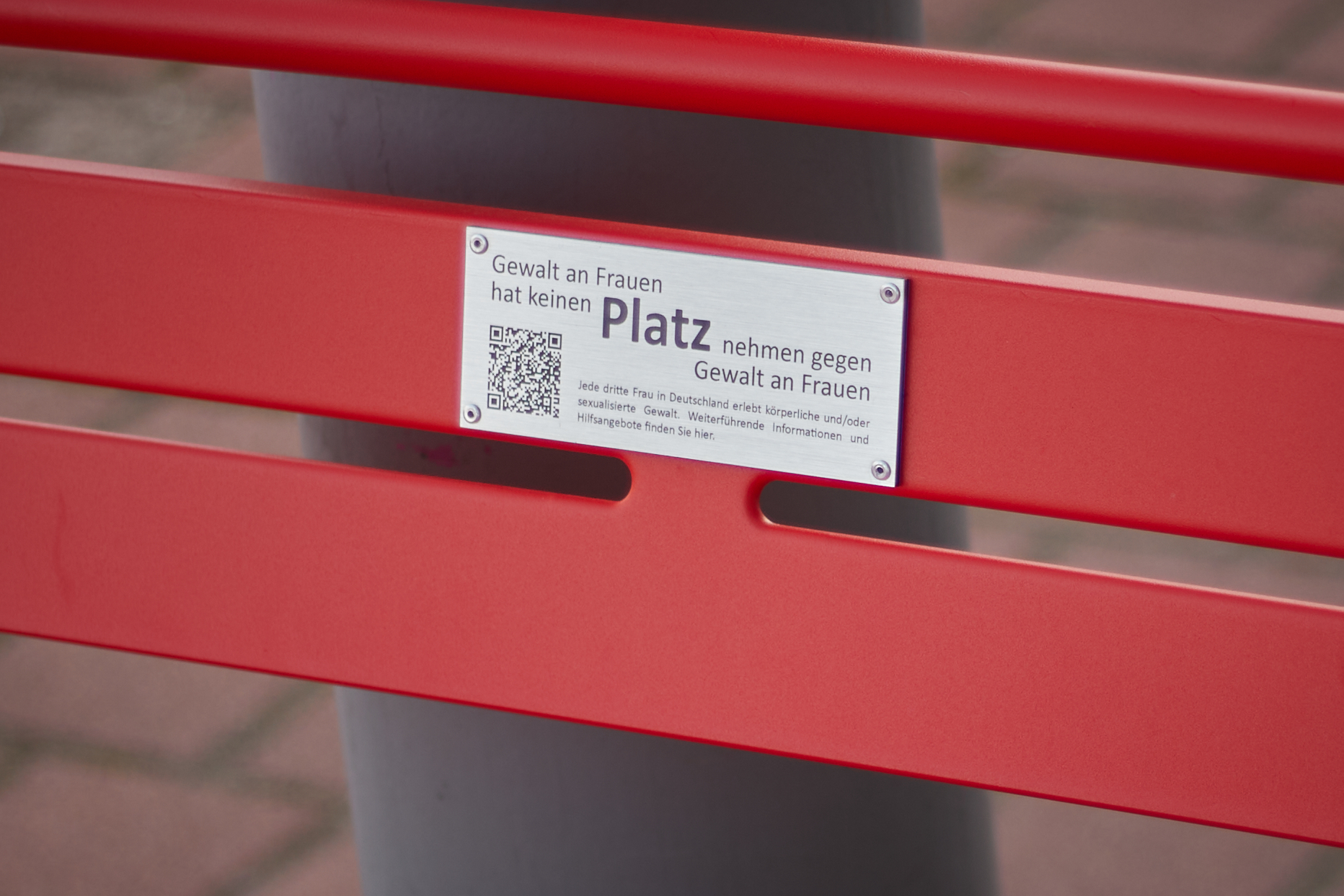 Nahaufnahme der Infoplakatte zur Aktion Rote Bank. Metallschild mit Text und QR-Code angebracht auf roten Holzpaneelen