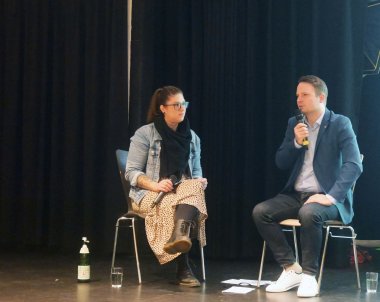 Jugendbeteiligung KGS Bürgermeister-Talk Zwei Personen sitzen auf Stühlen auf einer Bühne vor einem dunklen Hintergrund. Sie unterhalten sich.
