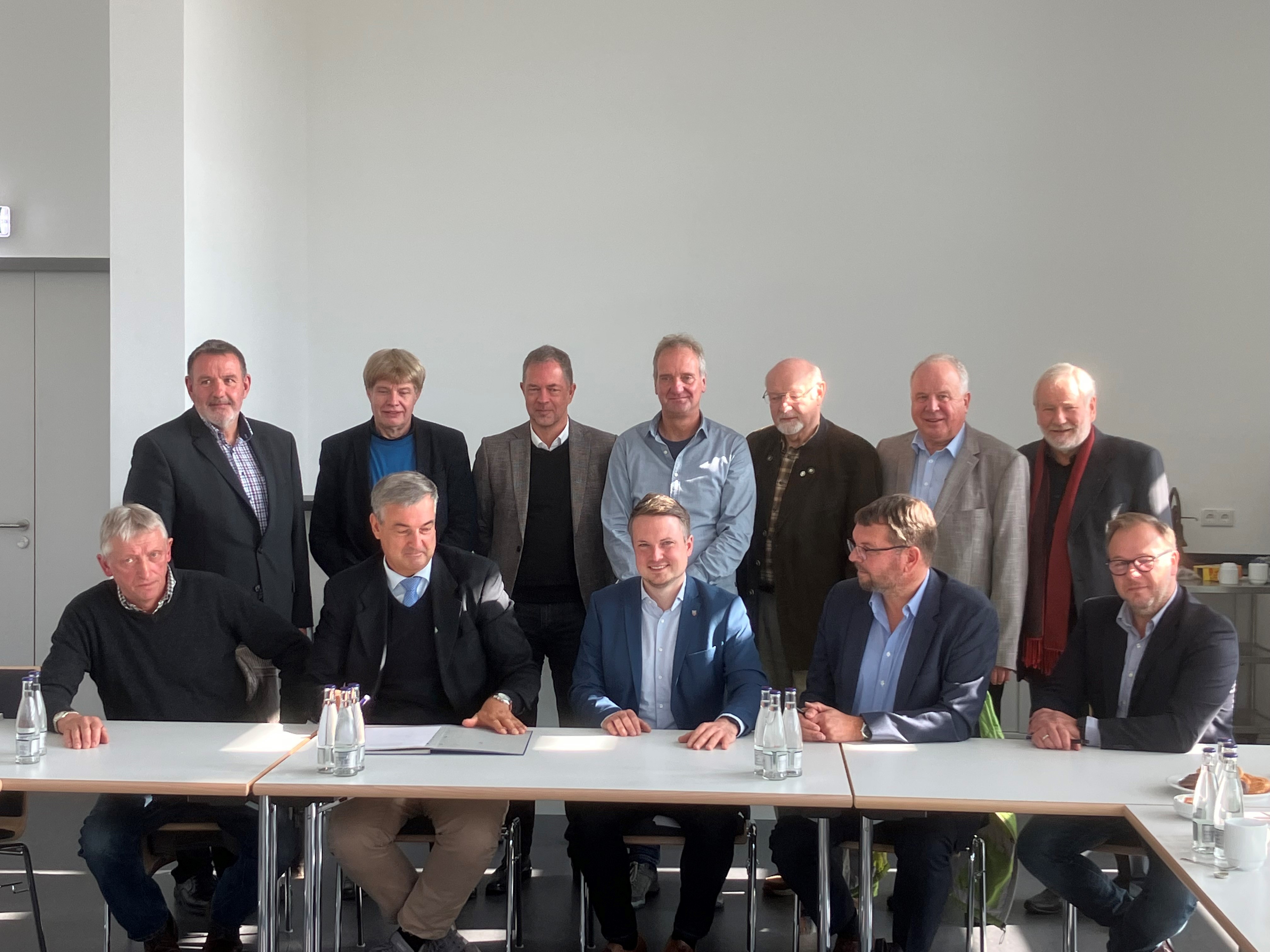 Gruppenbild mit 12 Personen, die in zwei Reihen hinter einem Tisch sitzen bzw. stehen. Vorne mittig sitzt der Bürgermeister, alle lächeln in die Kamera.