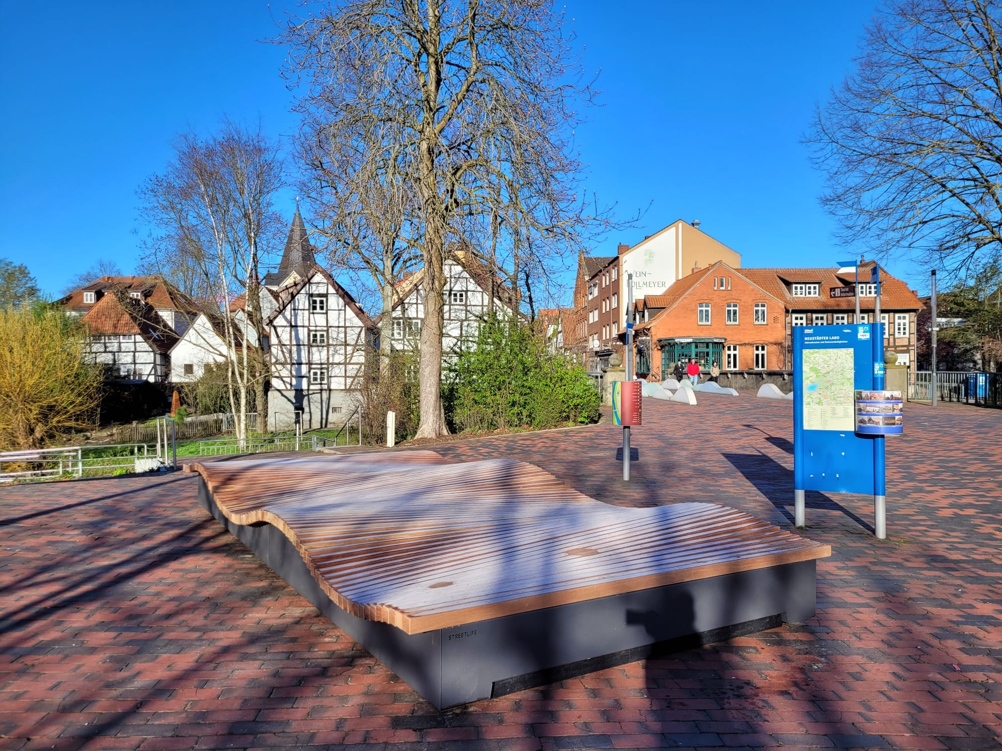 Auf dem Platz zwischen den Brücken steht ein neues Sitzmöbel. die große hölzerne Sitzfläche ist in Wellenform angelegt. Die Sonne scheint, der Schatten der Bäume fällt auf das Sitzmöbel.