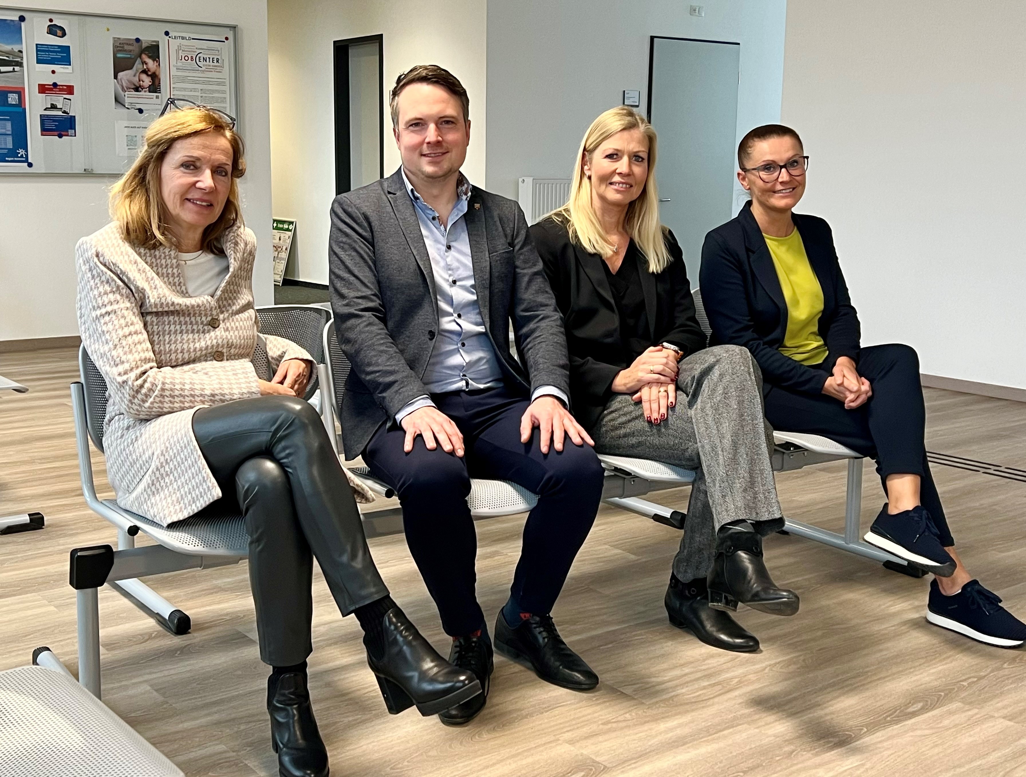 Vier Personen sitzen im Wartebreich des neuen JobCenters auf einer Bank. Alle Lächeln in die Kamera