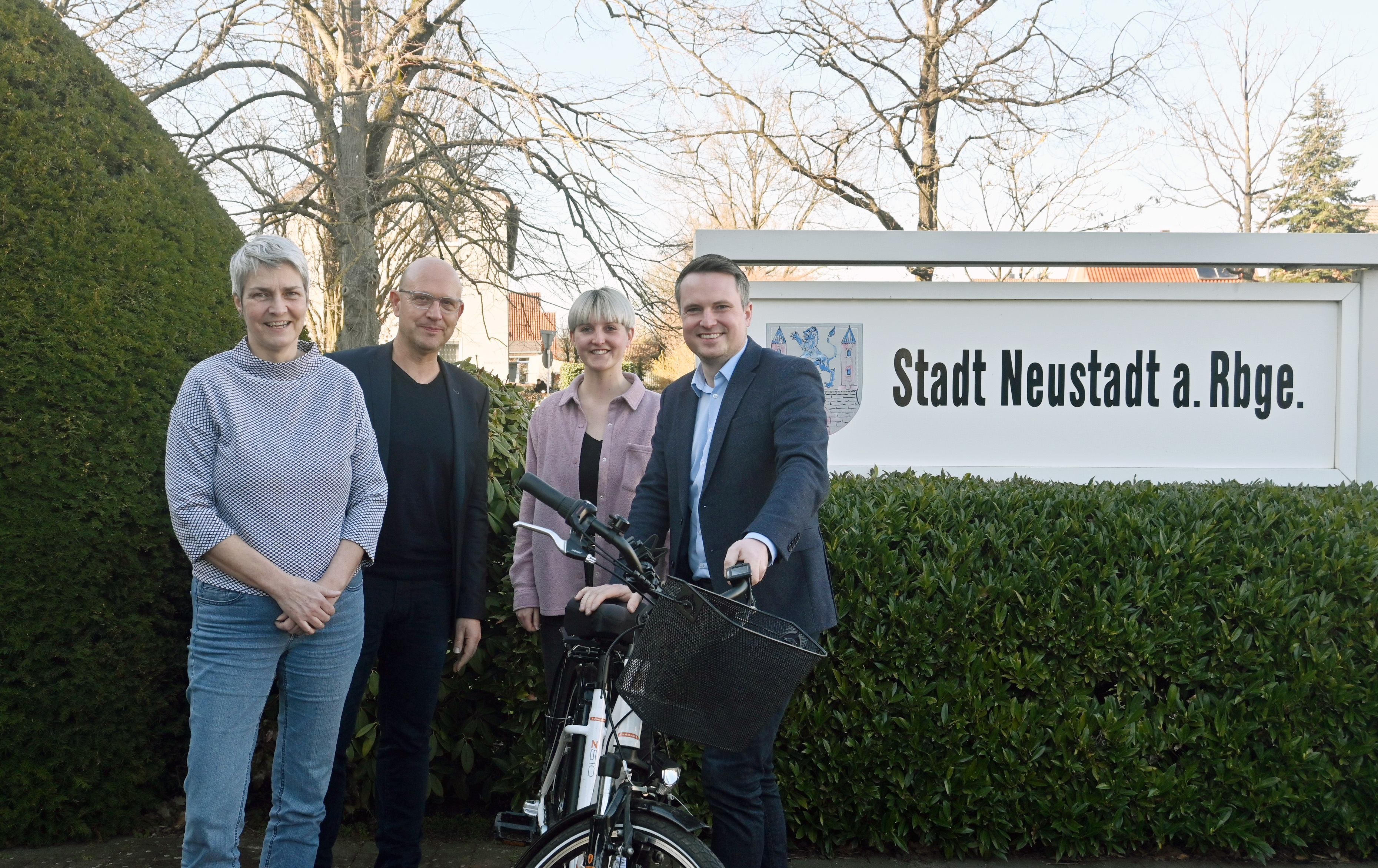 4 Personen stehen draußen vor einem Schild mit dem Wappen der Stadt Neustadt und der Aufschrift "Stadt neustadt a. Rbge.". Alle lächeln in die Kamera. Bürgermeister Dominic Herbst (links) hält ein weißes Fahrrad.