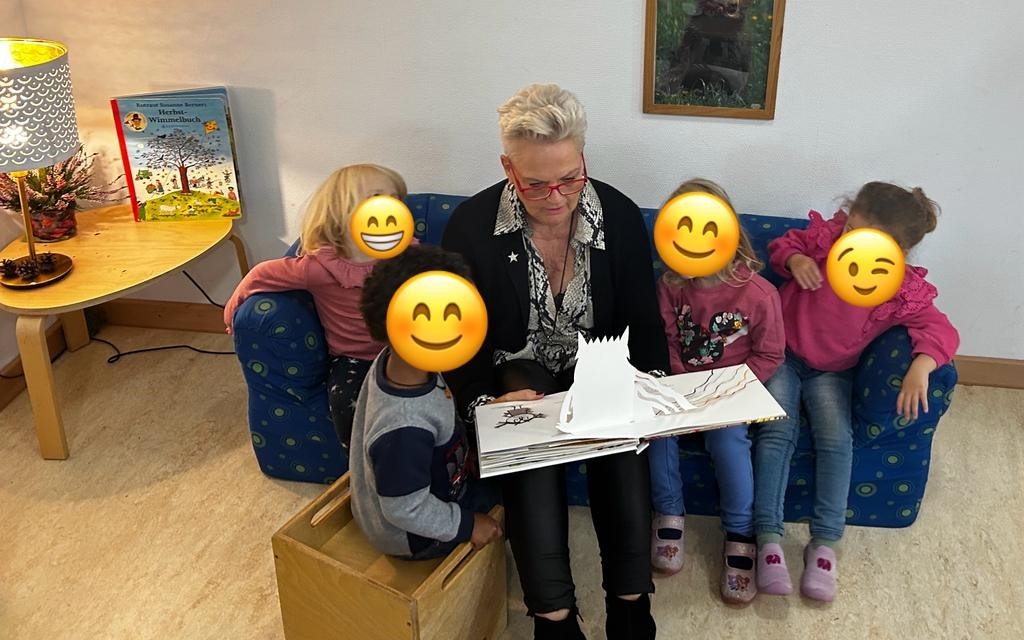 Eine Frau sitzt auf einem Sofa, um sie herum vier Kinder. Alle schauen in ein Buch.