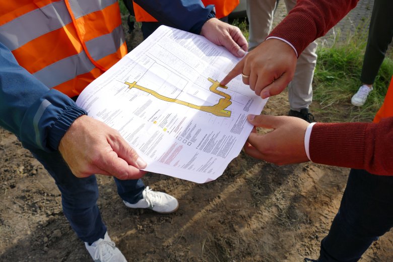 Nahaufnahme einer Planskizze - dort ist der zukünftige Brücke n- und Straßenverlauf in gelb markiert dargestellt. Zwei Hände halten den Plan, ein weiteres Paar hände zeigt auf den Plan