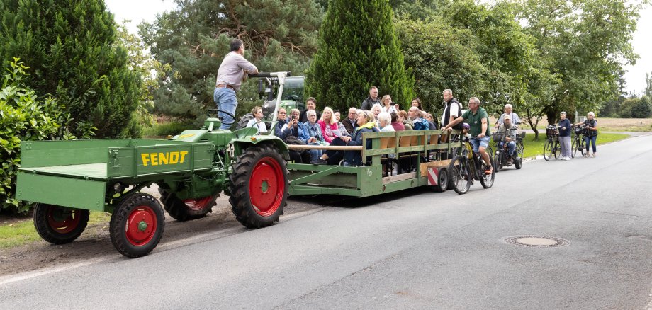 Besuch der Bewertungskommission in Bevensen zum 28. Regionswettbewerb Unser Dorf hat Zukunft Blick auf einen Trecker mit Anhänger auf dem zahlreiche Menschen sitzen. Sie werden von eingien Radfahrenden begleitet.