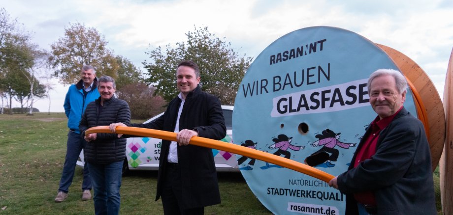 Vier Männer halten ein Glasfaserkabel. Sie stehen vor einer großen Kabeltrommel mit dem Schriftzug Rasannt. Wir bauen Glasfaser.