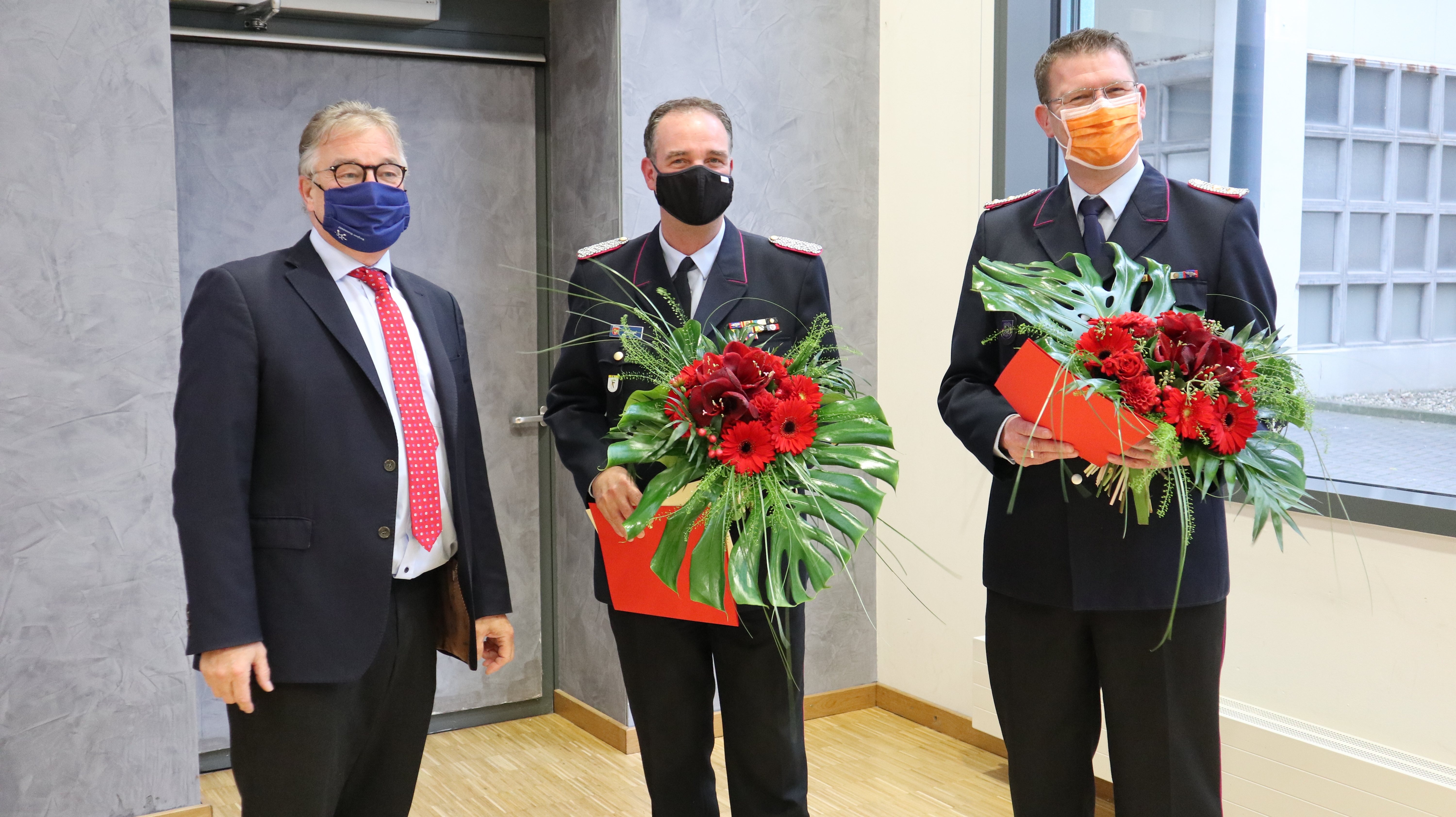 Drei Männer stehen nebeneinander. Die Herren mittig und links sind von der Feuerwehr und halten Blumensträuße in der Hand. Alle drei Tragen einen Mund-Nasen-Schutz.