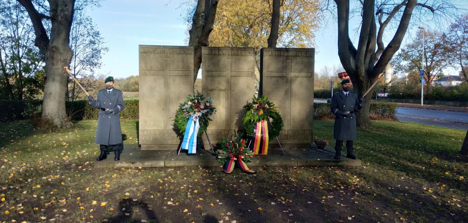 Zwei Soldaten mit Fackeln stehen neben dem steinernen Mahnmal. Es sind drei Trauerkränze niedergelegt.