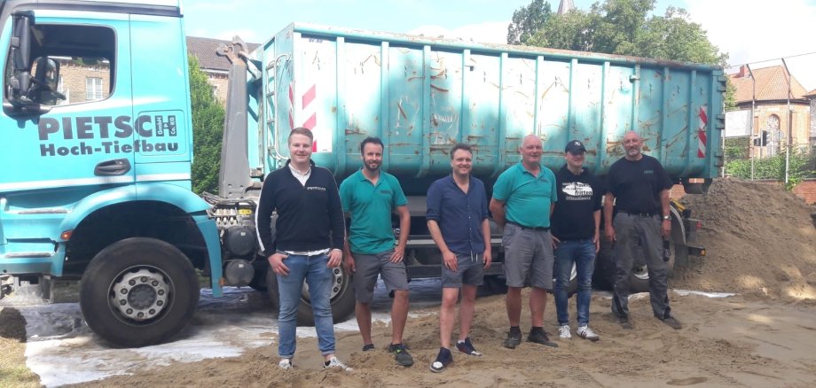 6 Männer stehen auf Sand vor einem großen Kipplaster und lächeln in die Kamera.