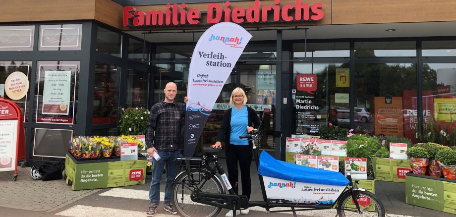 Zwei Personen stehen mit dem lastenrad Hannah und einer BEachflag vor dem Eingang des Rewew-Marktes an der Siemensstraße und lächeln in die Kamera