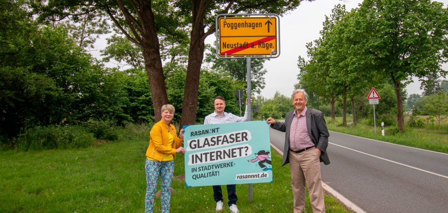 Drei Personen stehen mit einem Werbebanner von Rasannnt in den Händen vor dem Ortseingangsschild in Poggehagen. Im Hintergrund ist eine Straße und Bäume zu sehen, 