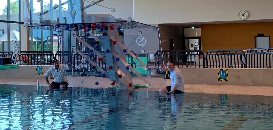 Zwei Männer sitze links und rechts neben der neuen Kletterwand am Schwimmbeckenrand, mit den Beinen im Wasser und lächeln in die Kamera.