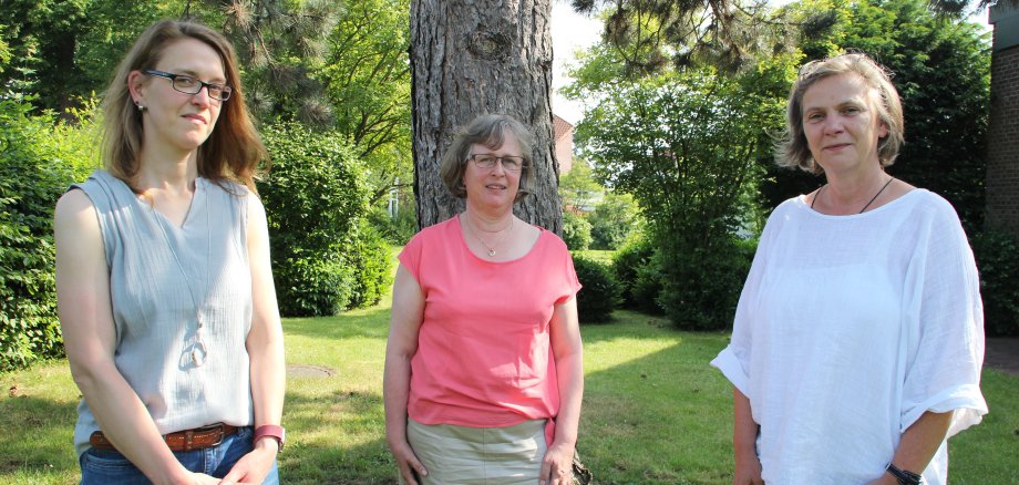 Drei Frauen stehen vor einem Baum auf einer Grünfläche und schauen in die Kamera.