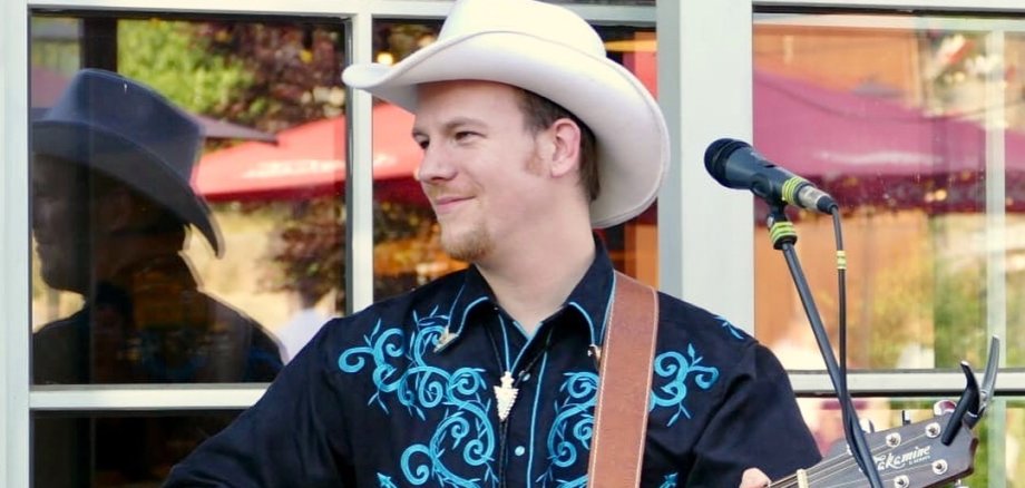Ein Country-Sänger mit Cowboyhut und Gitarre spielt Musik. Rechts neben ihm steht ein Mikrophonständer, er schaut nach links zur Seite