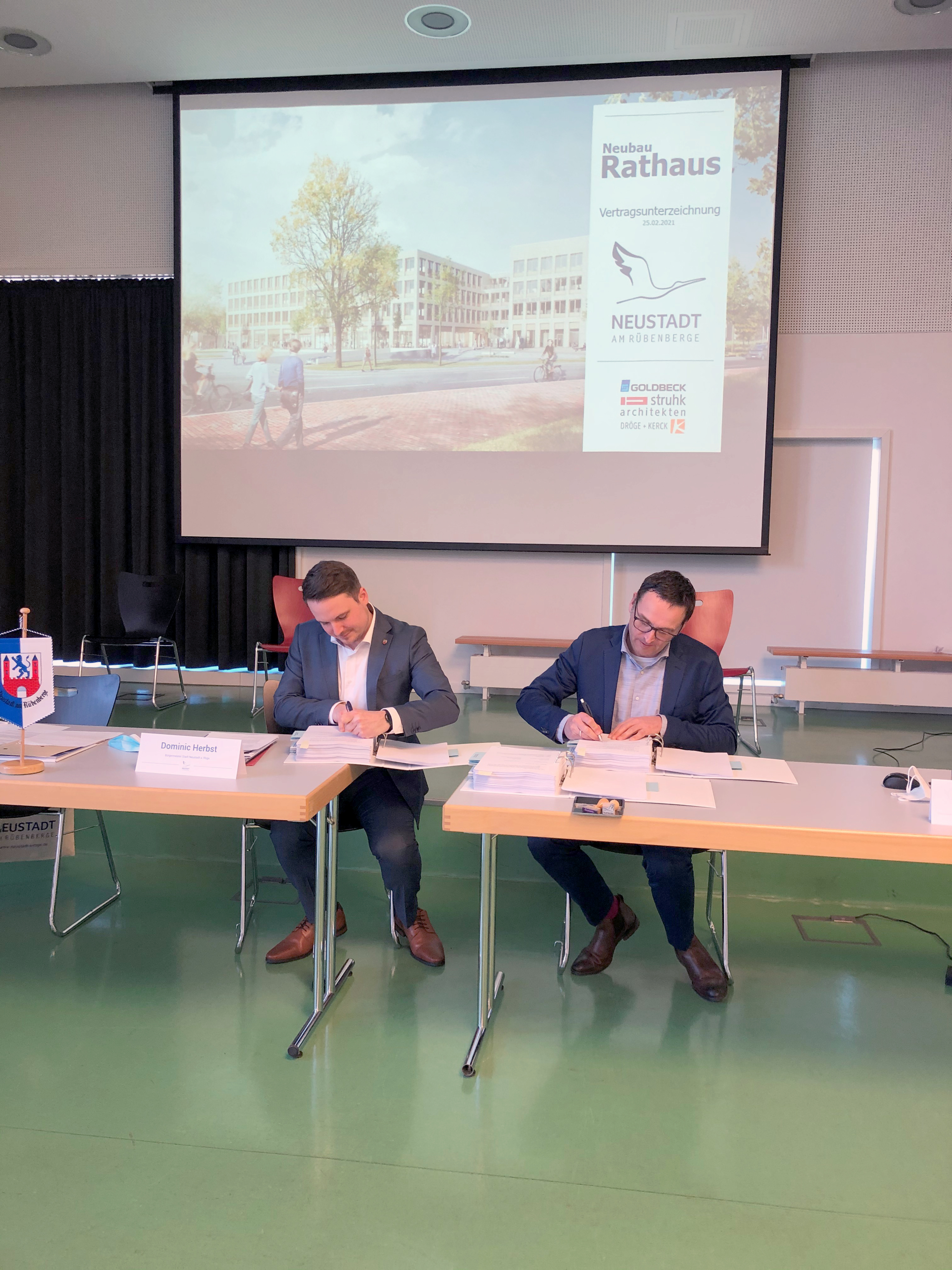 Zwei Männer sitzen an einem Tisch und unterschreiben Verträge. Im Hintergrund ist eine Leinwand, auf der das neue Rathaus zu sehen ist.