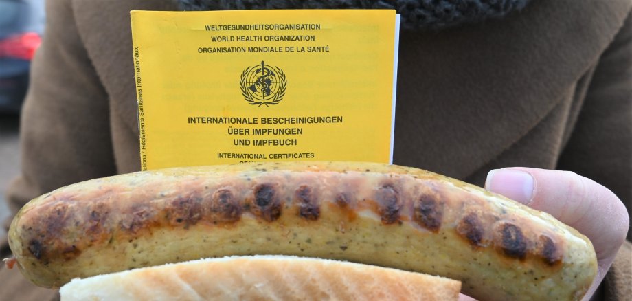 Eine Person hält eine Bratwurst in der Hand, dahinter ist ein gelber Impfpass zu sehen. 