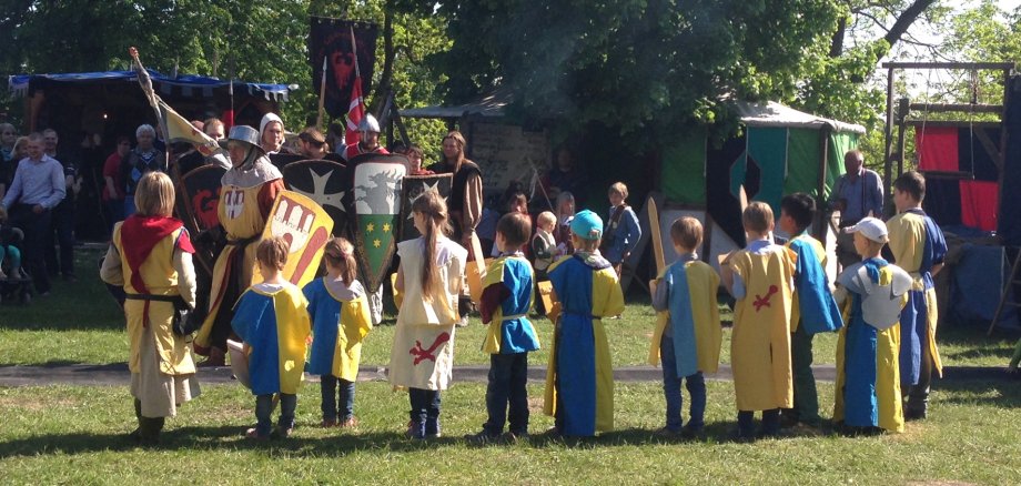 Gruppe von Kindern in Knappenköstümen steht auf einer grünen Wiese. Im Hintergrund sind Mittealterzelte und Ritter zu sehen.