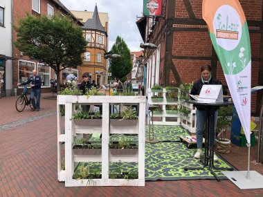 Blick auf den Stand zum mobilen Schaugarten. Links steht ein aus weißen Paletten gebautes kleines Planzbeet, rechts der Infotisch. Der Stand steht in der Fußgänerzone in Neustadt.