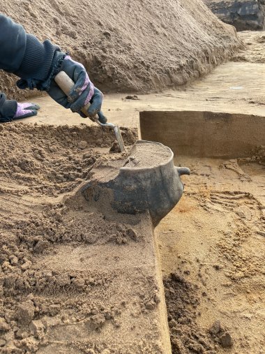 Nahaufnahme von einer Ausgrabung. Zwei Hände holen mit einer kleinen Schaufel Sand aus einer alterrümlichen Urne.