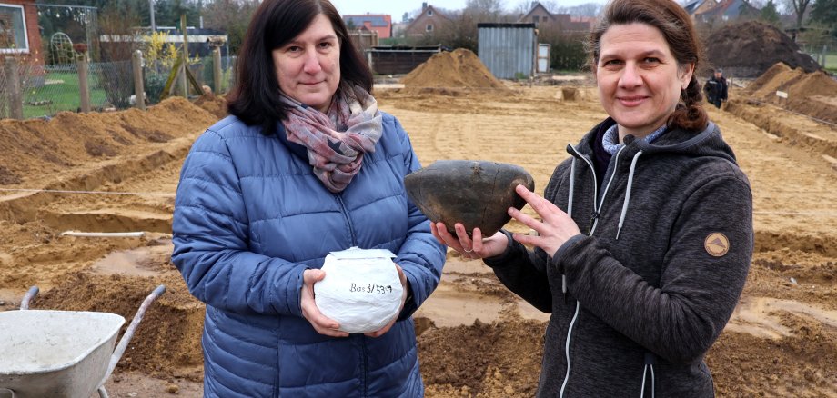 Die beiden Frauen stehen vor der Ausgrabunggstelle in Basse und halten je einen Fund in den Händen. Beide blicken in die Kamera.