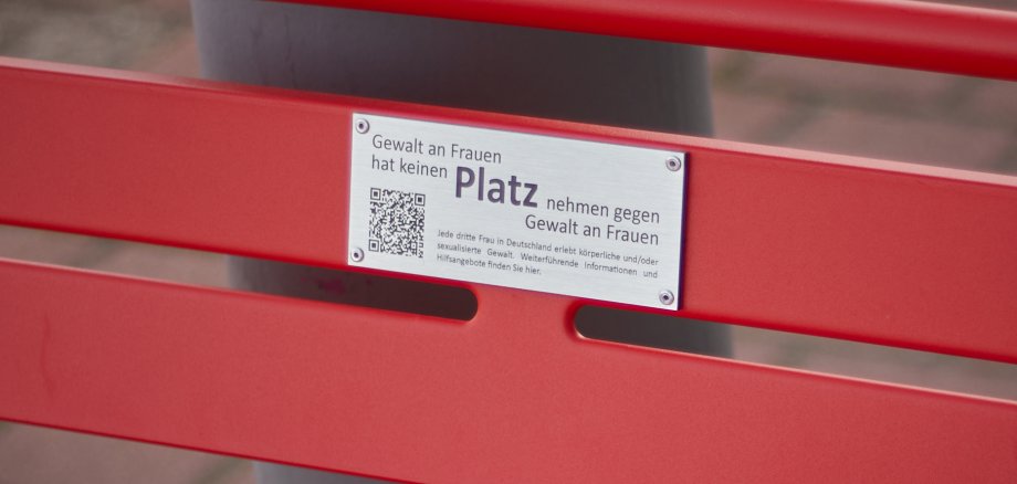 Nahaufnahme der Infoplakatte zur Aktion Rote Bank. Metallschild mit Text und QR-Code angebracht auf roten Holzpaneelen