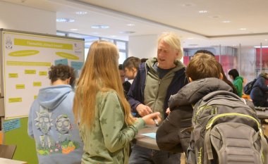 Drei Personen stehen an einem Stehtisch und sprechen. Im Hintergrund ist eine Stellwand mit Vorschlägen und Ideen zu sehen.