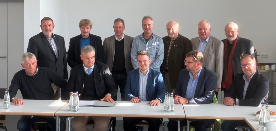 Gruppenbild mit 12 Personen, die in zwei Reihen hinter einem Tisch sitzen bzw. stehen. Vorne mittig sitzt der Bürgermeister, alle lächeln in die Kamera.