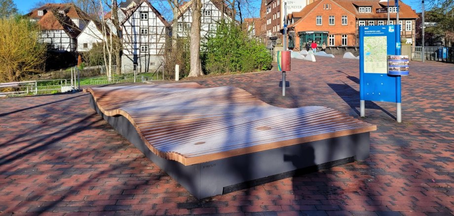 Auf dem Platz zwischen den Brücken steht ein neues Sitzmöbel. die große hölzerne Sitzfläche ist in Wellenform angelegt. Die Sonne scheint, der Schatten der Bäume fällt auf das Sitzmöbel.