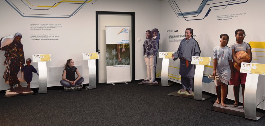 Aufnahme Teil der Klimafluchtausstellung. Lebensgroße pappfiguren verschiedener betroffener Personen stehen in einem Raum an der Wand, naben stehen Aufsteller mit Infotafeln.