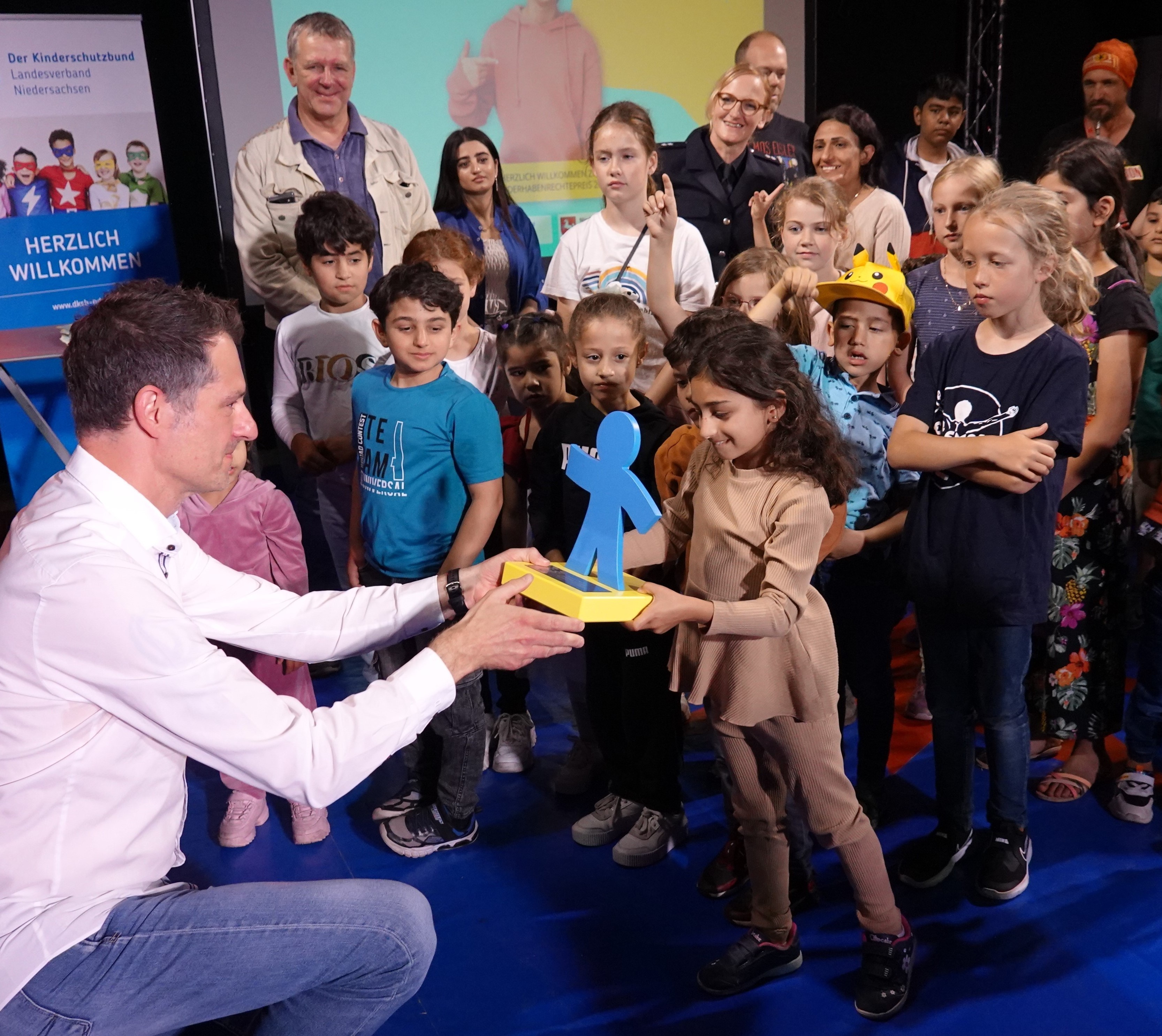 Pressefoto Neustädter Netzwerk erhält Preis Eine Gruppe von Menschen steht auf einer Bühne. im Vordergrund kniet ein Mann und übrgibt einen Preis in Form einer blauen Figur an ein kleines Mädchen.