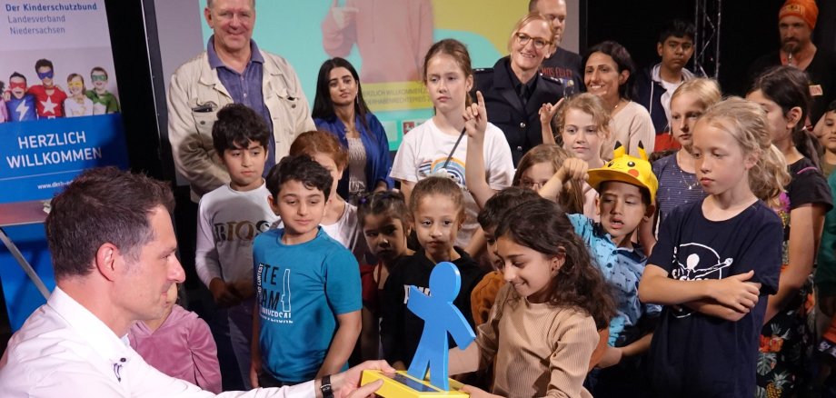 Eine Gruppe von Menschen steht auf einer Bühne. im Vordergrund kniet ein Mann und übrgibt einen Preis in Form einer blauen Figur an ein kleines Mädchen.