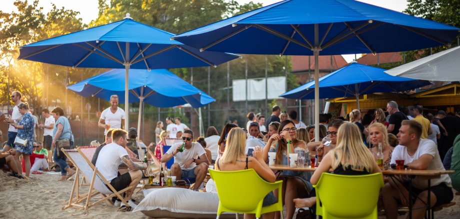 Blick auf den Betrieb des Citybeaches. Menschen sitzen an einem Sommerabend auf Liegestühlen unter Sonnenschirmen auf der Sandfläche vor dem Schloss und genießen Essen und Getränke.