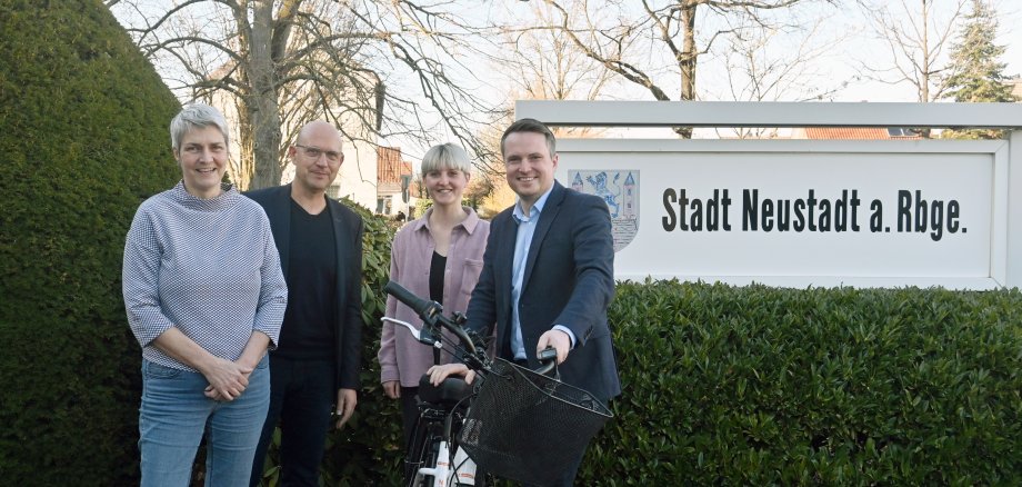 4 Personen stehen draußen vor einem Schild mit dem Wappen der Stadt Neustadt und der Aufschrift "Stadt neustadt a. Rbge.". Alle lächeln in die Kamera. Bürgermeister Dominic Herbst (links) hält ein weißes Fahrrad.