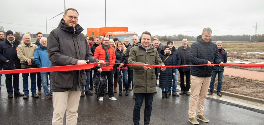 (v.l.) Thomas Reimann, Projektleiter für das Gebiet und Prokurist im Ideenstadtwerke Konzern, Bürgermeister Dominic Herbst und Ideenstadtwerke-Geschäftsführer Dieter Lindauer schneiden das symbolische rote Band zur Einweihung des neuen Gewerbegebiets durch. Im Hintergrund freuen sich Aufsichtsratsmitglieder, Käuferinnen und Käufer, beteiligte Unternehmen sowie Mitarbeiterinnen und Mitarbeiter der Stadt sowie der Ideenstadtwerke.