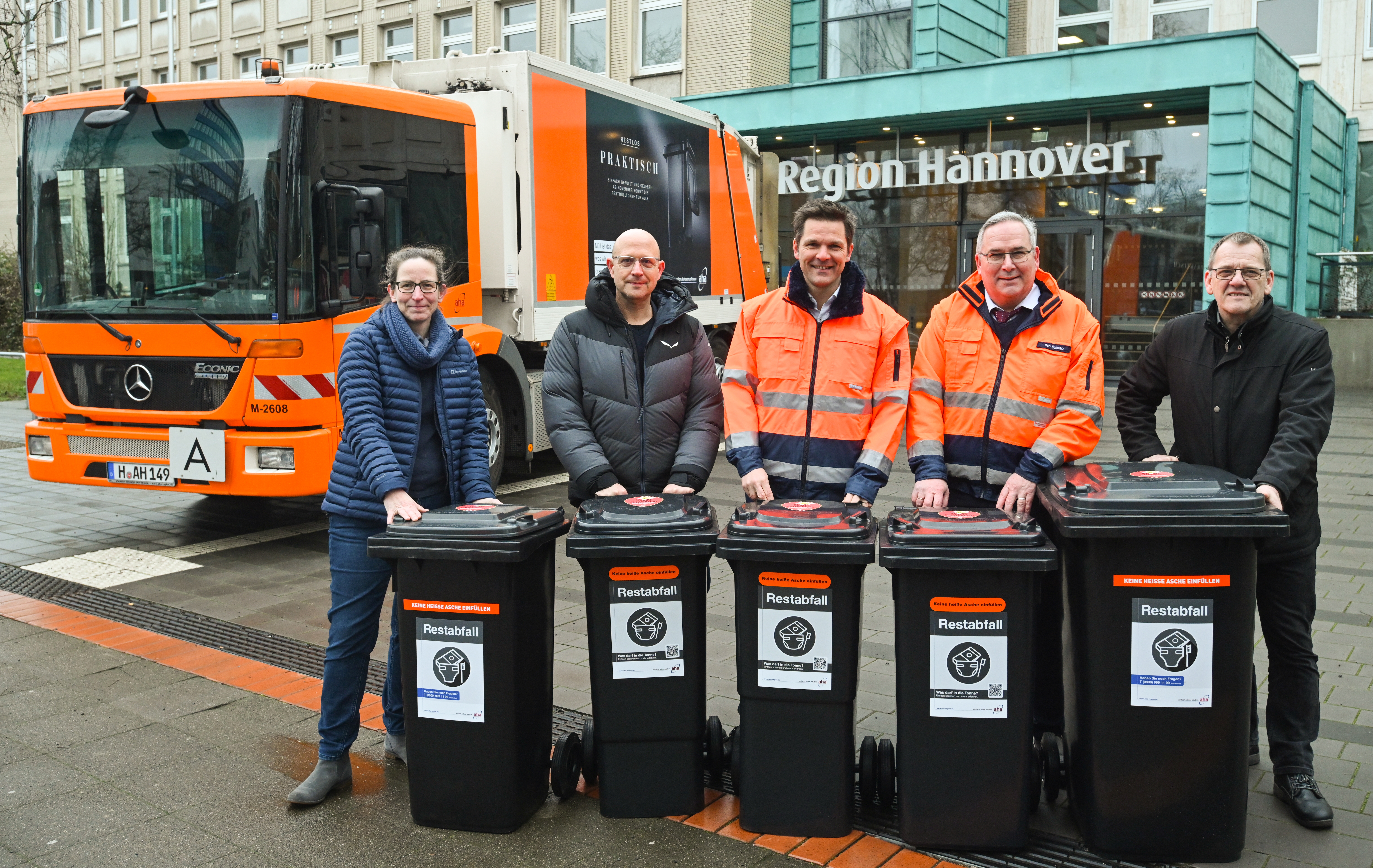 fünf Personen stehen mit jeweils einer grauen Restmülltonne vor dem Regionshaus in Hannover. Im Hintergrund ist ein Müllwagen zu sehen.