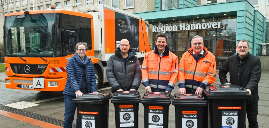 fünf Personen stehen mit jeweils einer grauen Restmülltonne vor dem Regionshaus in Hannover. Im Hintergrund ist ein Müllwagen zu sehen.