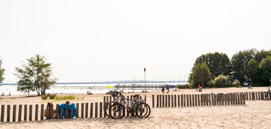 Blick auf den Badestrand am Steinhuder meer in mardorf. Zwei Fahrräder stehen angeschlossen an Holpollern. im Hintergrund ist das Steinhuder Meer sowie einzelne Badegäste erkennbar.