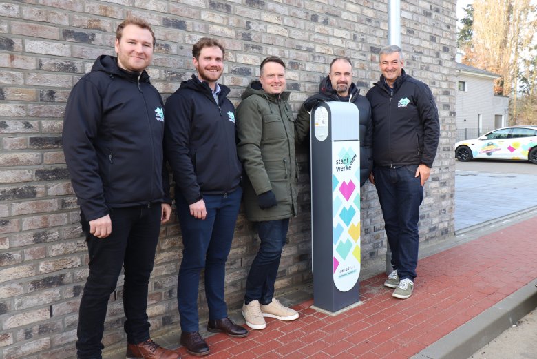 Fünf Personen stehen vor einer Hauswand und blicken in die Kamera. In der Mitte steht eine Ladesäule für E-Autos der Stadtwerke.