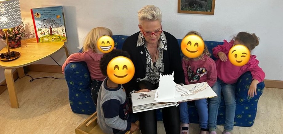 Eine Frau sitzt auf einem Sofa, um sie herum vier Kinder. Alle schauen in ein Buch.