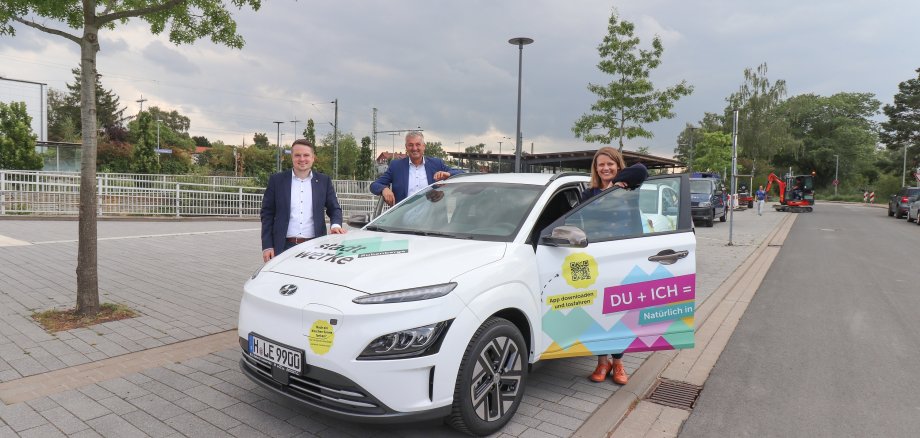 Blick auf die Front des E-Autos der Carsharing-Flotte. Das Auto ist bunt beklebt. Julia Willie Hamburg steht rechts hinter der geöffneten Fahrertür, Dieter Lindauer hinten links am Auto und Dominic Herbst vorn links neben dem Wagen. Alle lächeln in die Kamera.