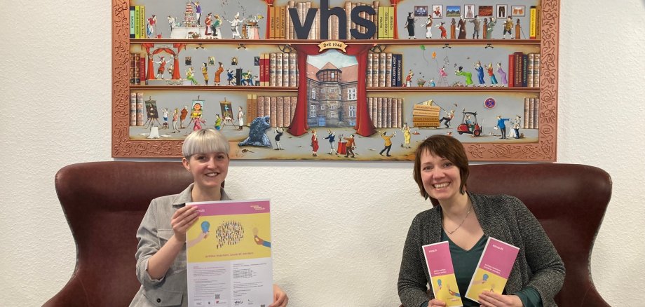 Wendy Pfeil (links) und Ulrike Hoffmann (rechts) sitzen auf zwei großen brauinen Sesseln. Wendy Pfeil hält ein Plakat in den Händen, Ulrike Hoffmann zwei Infoflyer. Beide lächeln in die Kamera.