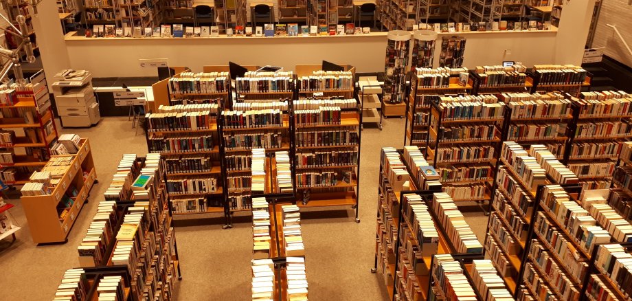 Lesesaal der Stadtbibliothek. Es stehen viele Regale voller Bücher in einem großem Raum.