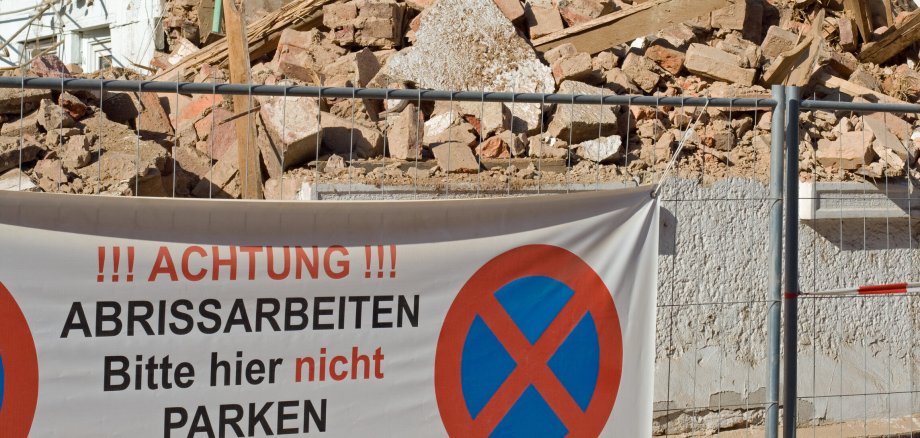 Symbolbild Abrissarbeiten Blick auf eine Baustelle mit einem Haufen Schutt. Davor ein Bauzaun mit einem Banner. Darauf ein Parkverbotsschild und der Schriftzug Achtung Abrissarbeiten