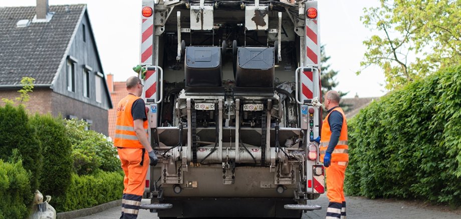 Aufnahme vom hinteren Teil eines Müllwagens, der gerade zwei Restmülltonnen leert. Links und rechts daneben stehen jeweils ein Mann in der Arbeitskleidung der Müllabfuhr