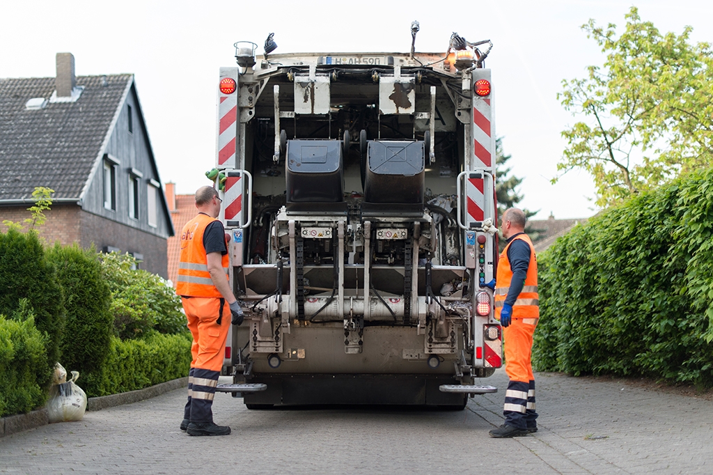 Aufnahme vom hinteren Teil eines Müllwagens, der gerade zwei Restmülltonnen leert. Links und rechts daneben stehen jeweils ein Mann in der Arbeitskleidung der Müllabfuhr