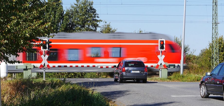 Symbolbild Bahnübergang Zu sehen ist eine geschlossene Bahnschranke. Davor wartet ein Auto, es fährt ein Zug vorbei