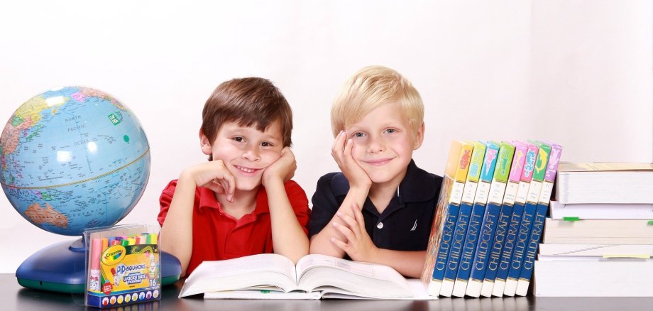 Zwei Jungs im Grundschulalter sitze an einem Tisch und lächeln in die Kamera. AUf dem Tisch steht ein Globus, Schulutensilien und ein Stapel mit Büchern. 
