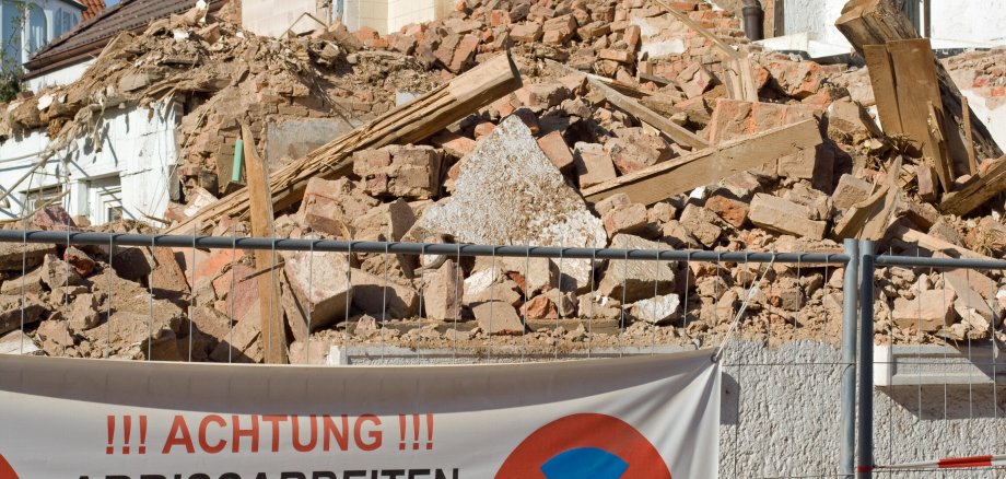 Blick auf eine Baustelle mit einem Haufen Schutt. Davor ein Bauzaun mit einem Banner. Darauf ein Parkverbotsschild und der Schriftzug Achtung Abrissarbeiten