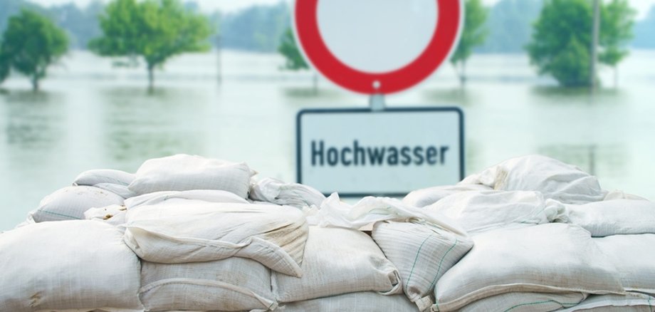 Schild mit der Aufschrift Hochwasser steht hinter gestapelten Sandsäcken. Im Hintergrund ist Wasser zu sehen. 