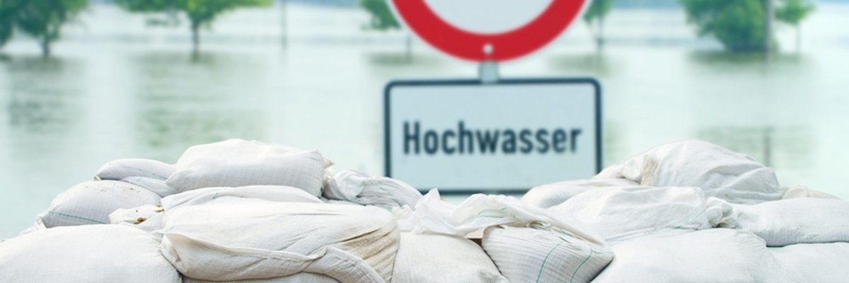 Symbolbild Hochwasser Schild mit der Aufschrift Hochwasser steht hinter gestapelten Sandsäcken. Im Hintergrund ist Wasser zu sehen.